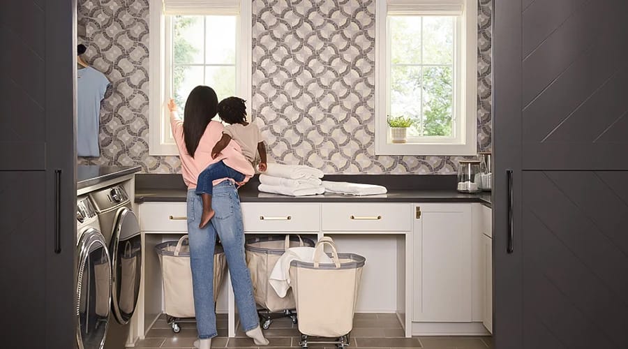 A woman holding a child in a quality laundry room with patterned wallpaper and modern appliances.