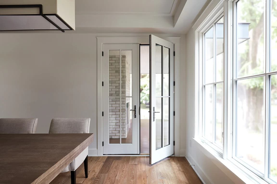 A white hinged door with glass panels opens from a bright dining room with wood floors and large windows.