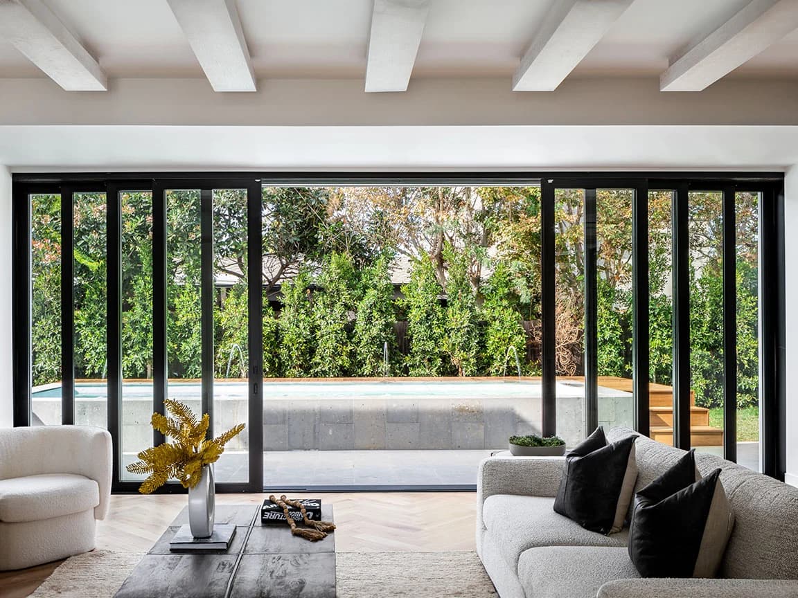 A modern living room with multi-slide glass doors opening to a pool and lush garden view.