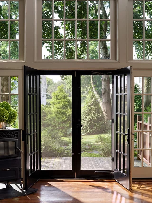 A black hinged door with glass panels opens to a lush garden, framed by large windows and wooden floors.