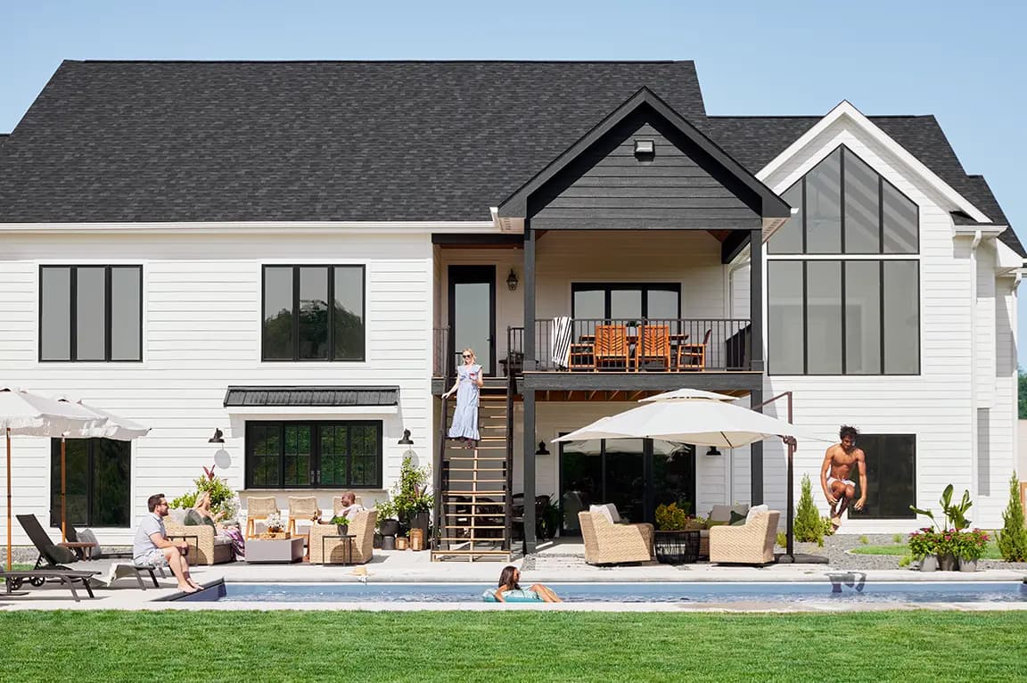 Two-story modern white farmhouse with black roof, backyard pool, patio seating and people relaxing outdoors