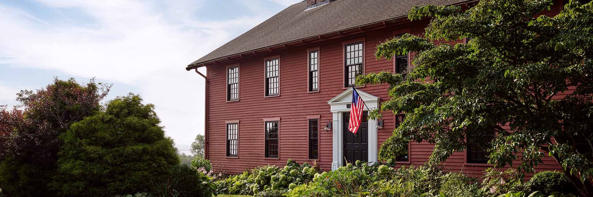 Red two-story colonial house with white trim, black front door and American flag, surrounded by lush garden and trees