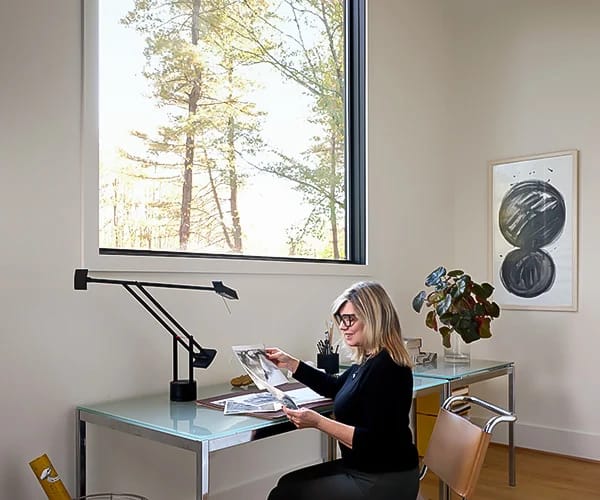 A woman reviews papers at a glass desk in a modern home office with a large window, ideal for sound performance.