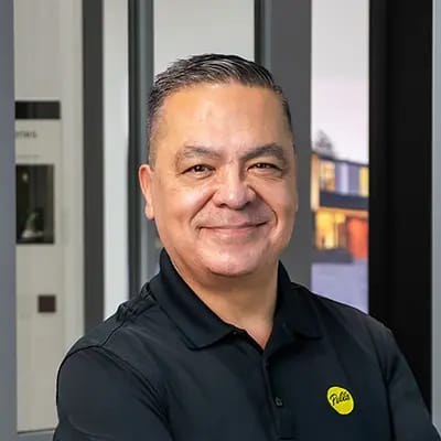 Smiling man in black polo shirt with yellow logo, arms crossed in a modern window showroom