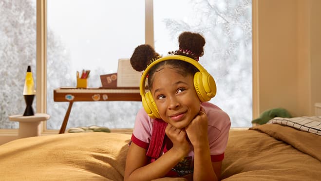 A small child wearing yellow headphones is dancing in front of a window wall while it is snowing outside.