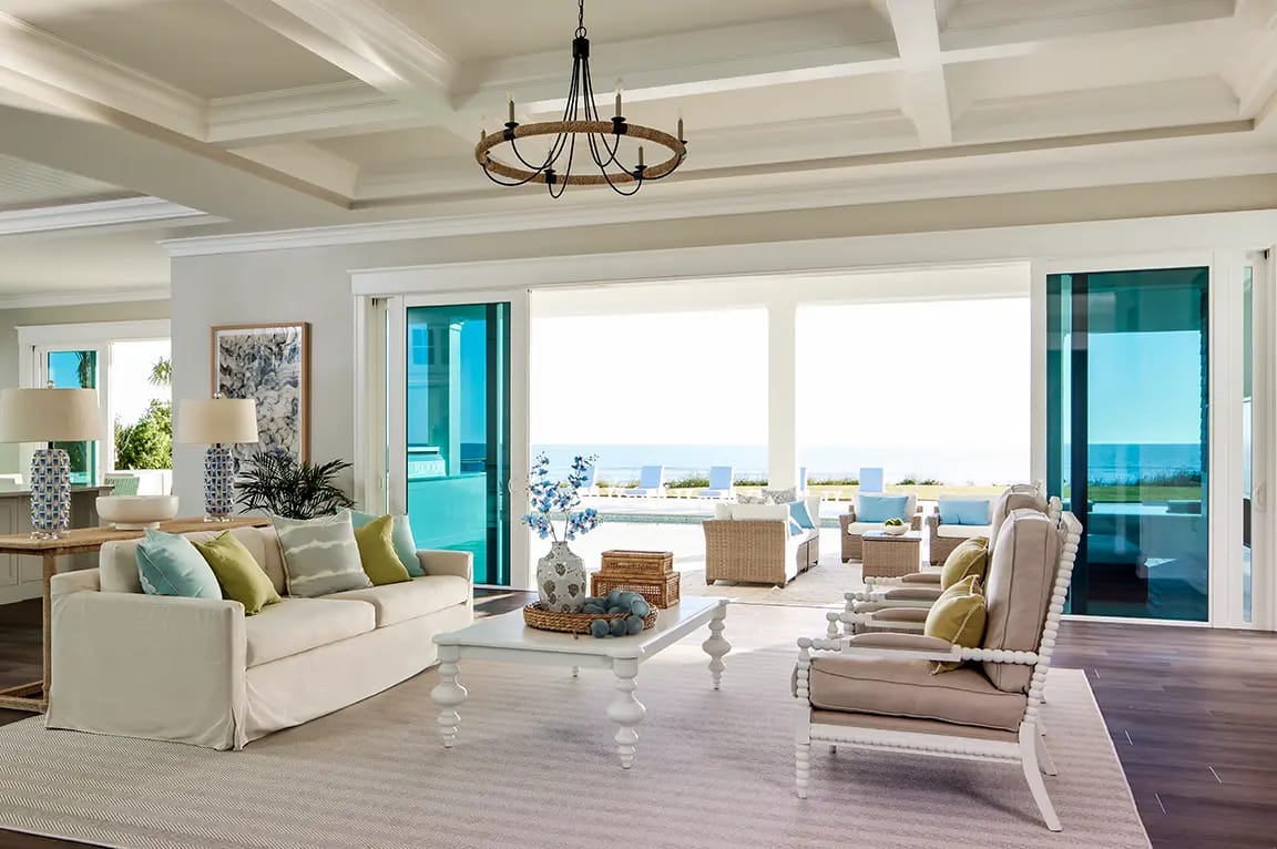 Beachfront coastal living room with neutral sofa, blue-green accents, white coffee table and sliding glass doors opening to ocean patio