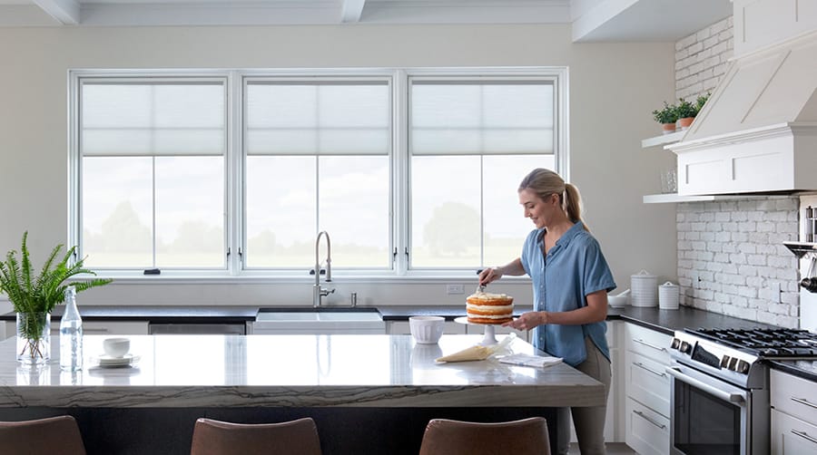 a woman frosts a cake in her kitchen in front of three pella casement windows featuring blinds-between-the-glass