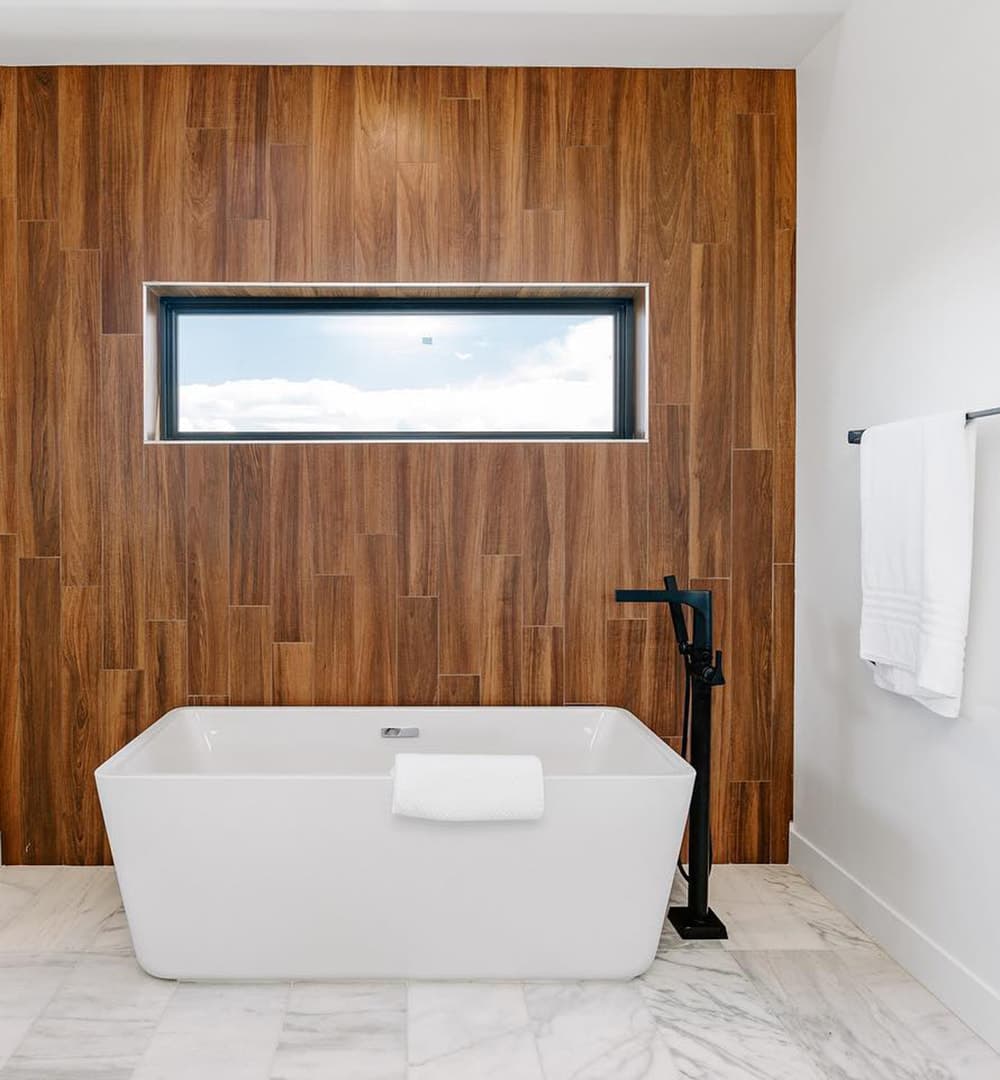 A wood accent wall features a rectangular picture window above a bathtub.