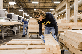 A woman wearing safety glasses is arranging a palette of fresh-cut wood pieces.
