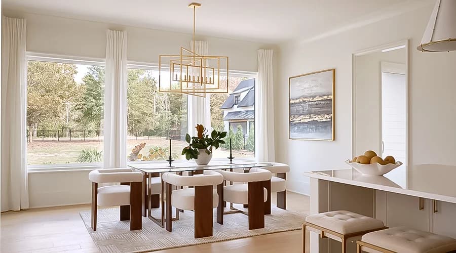 A modern dining room with large windows showing sunny weather and trees outside.