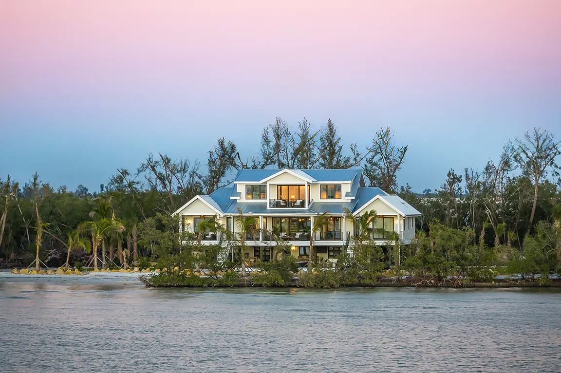 Luxury two-story waterfront home with blue metal roof and palm trees on a mangrove shoreline at sunset