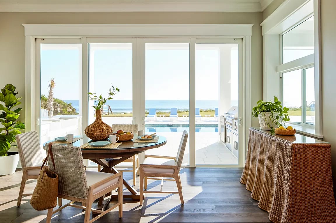 Sunlit coastal dining area with round wooden table, wicker chairs and rattan console, sliding glass doors opening to pool and ocean view