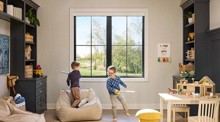 Two children perform playful activities with toys in a bright room featuring a large window and organized shelves.