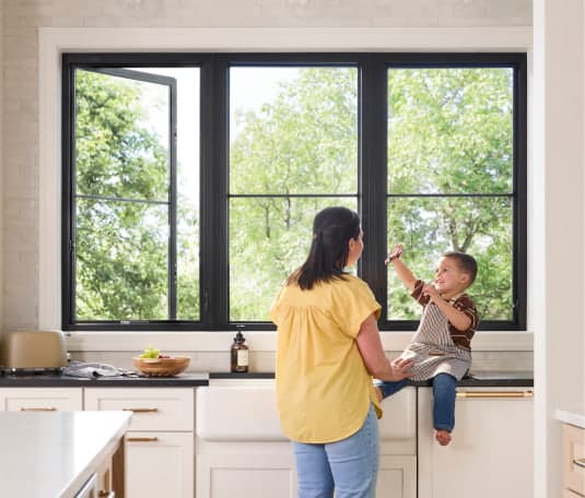 A mother and child interact in a bright kitchen with a large black-framed window overlooking green trees.