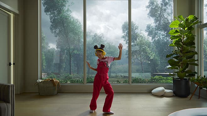 A young girl is wearing yellow headphones and pink overalls and dancing in front of a large multi-slide door while the wind is blowing outside.