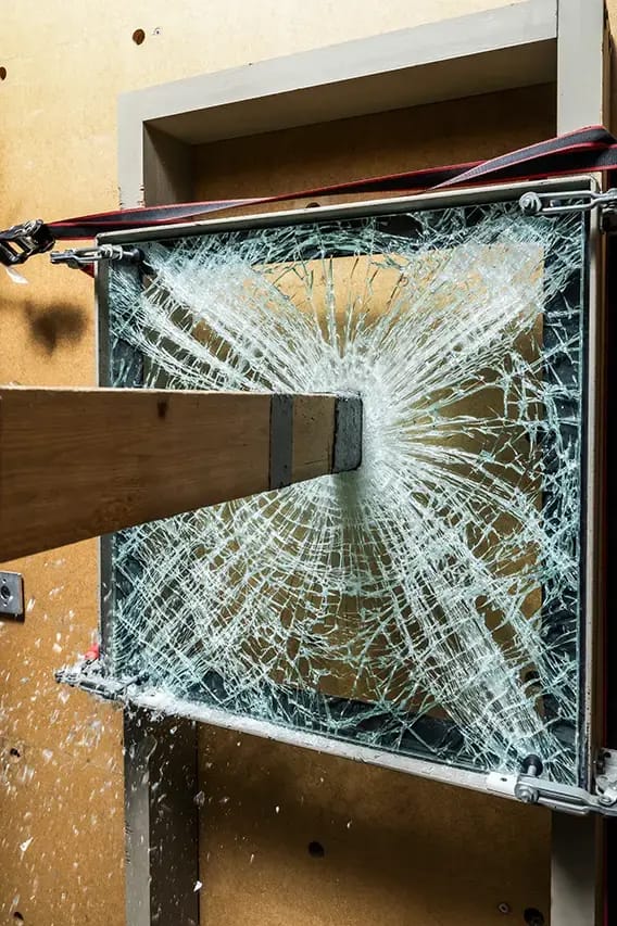 Shattered laminated safety glass panel with a wooden beam lodged at the center, spiderweb radial cracks from an impact test
