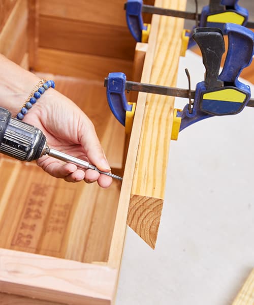 Two clamps are holding a board in place while a long screw is being fastened to securely attach to the window box.
