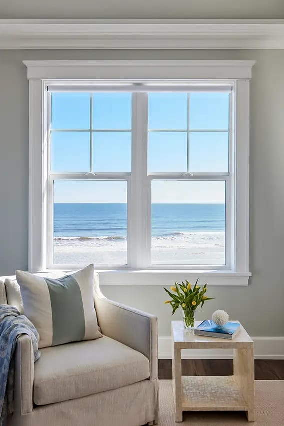 Ocean-view window in coastal living room overlooking blue ocean and sandy beach, beige armchair and side table with yellow tulips.