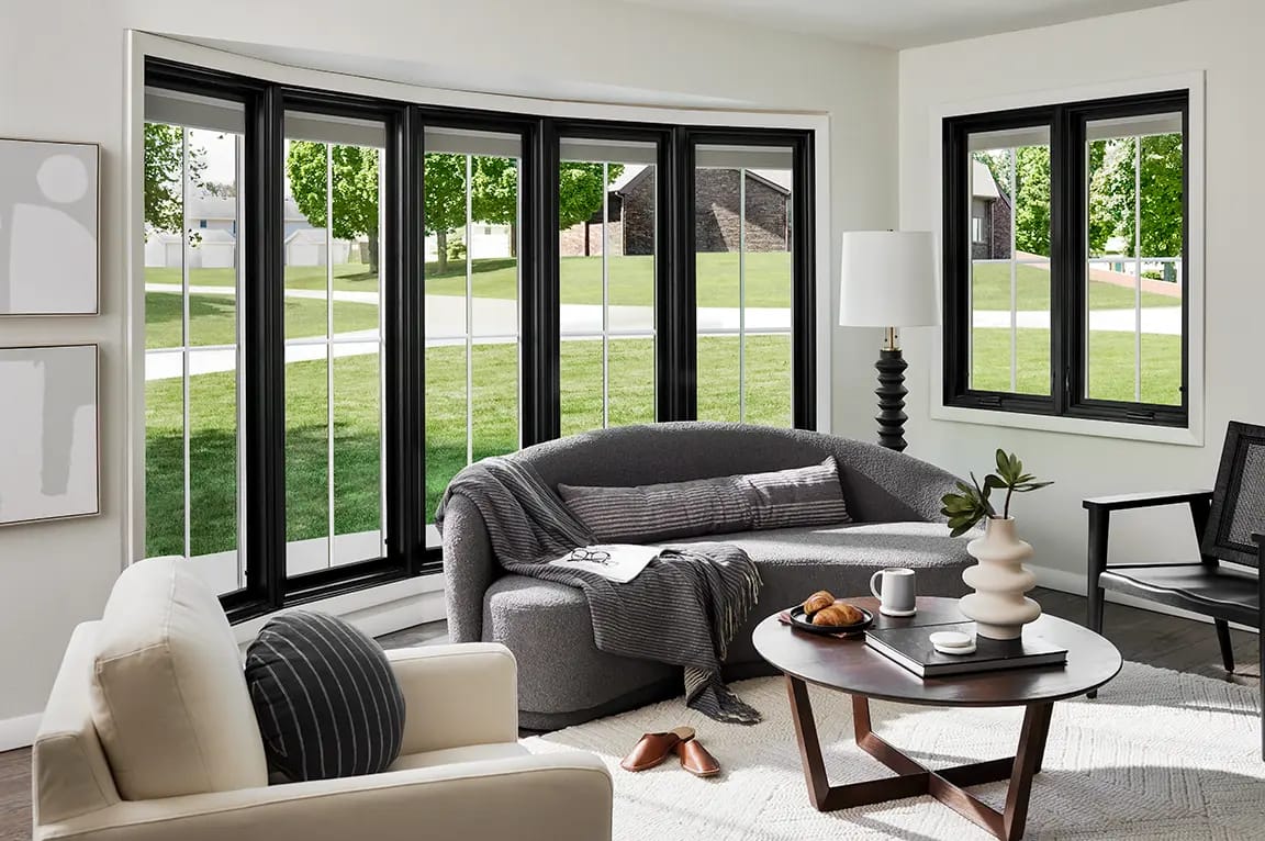 Modern living room with large black-framed bay windows, curved gray sofa, cream armchair, round wooden coffee table and neutral decor
