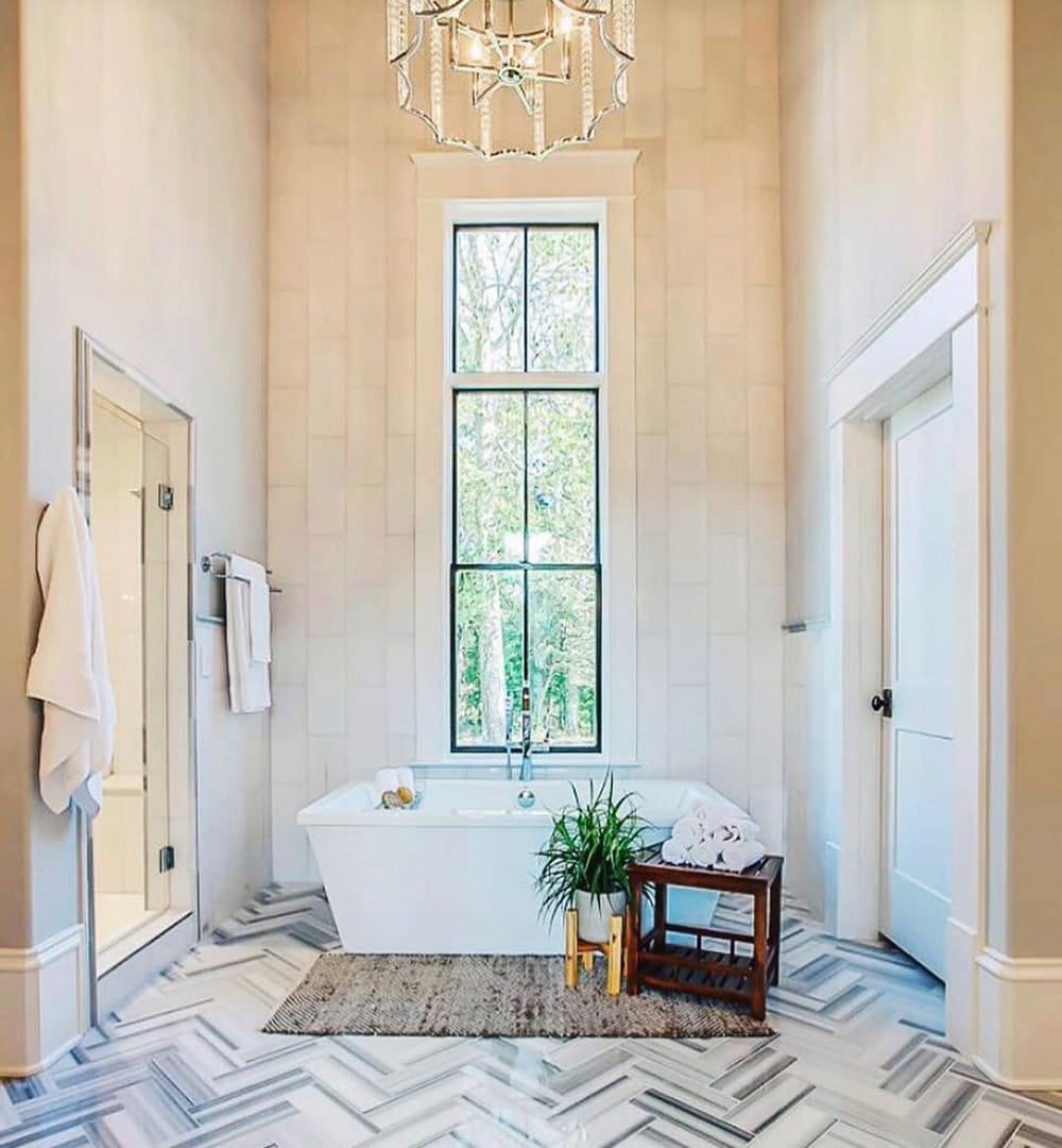 A modern and contemporary bathroom with high ceilings and a chandelier features a bathtub with a tall black window above.