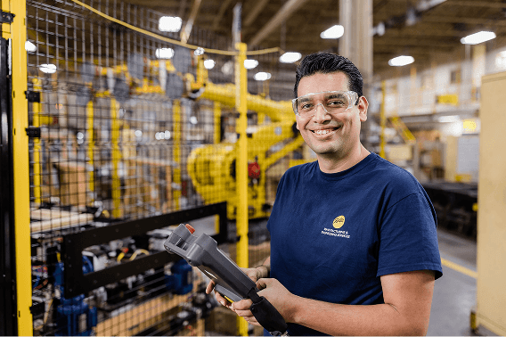 A man wearing a blue shirt is holding a robotic control while standing in a manufacturing plant.