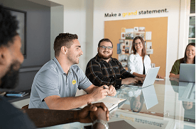 A group of people are smiling while sitting around a meeting table with their laptops